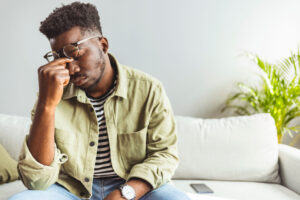 Man sitting on couch reaching under glasses to pinch bridge of nose.