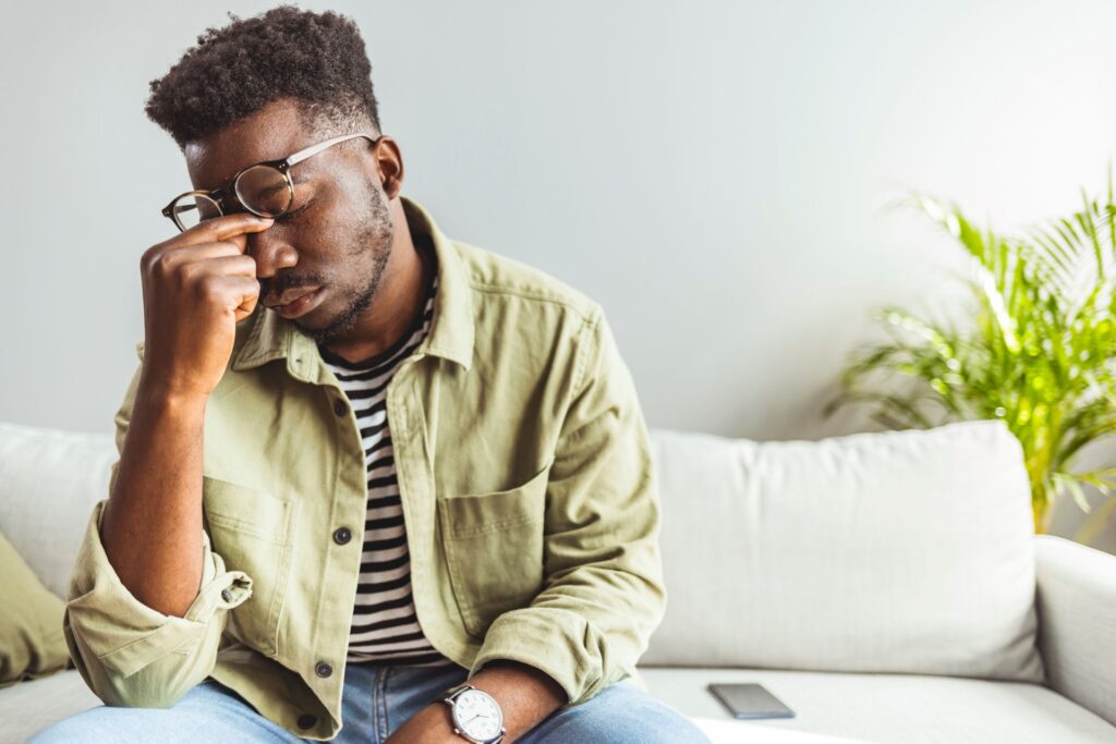 Man sitting on couch reaching under glasses to pinch bridge of nose.